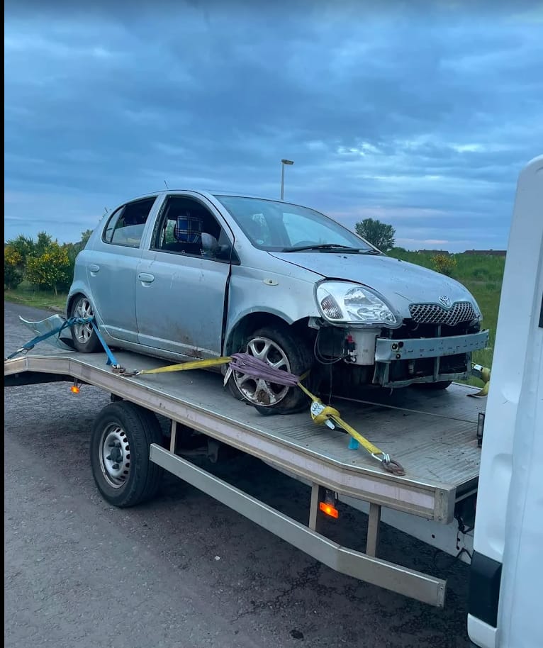 A silver Toyota Yaris with front-end accident damage loaded on a Speedy Recovery flatbed at dusk